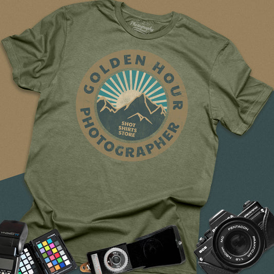 A military green t-shirt featuring a distressed circular vintage badge design with mountain peaks and sunburst rays, surrounded by the text "Golden Hour Photographer" written along the border.