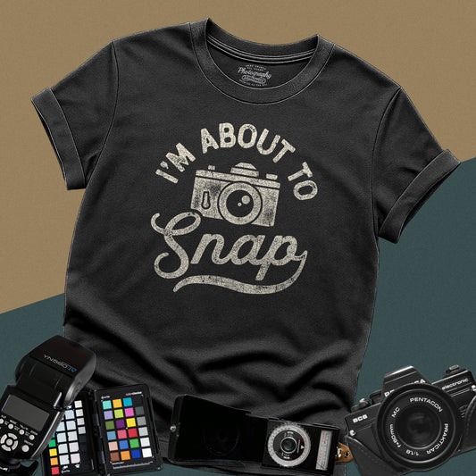 A black t-shirt featuring a distressed vintage camera graphic centered between the text "I'm about to" in a curved font and "Snap" written in retro script below.