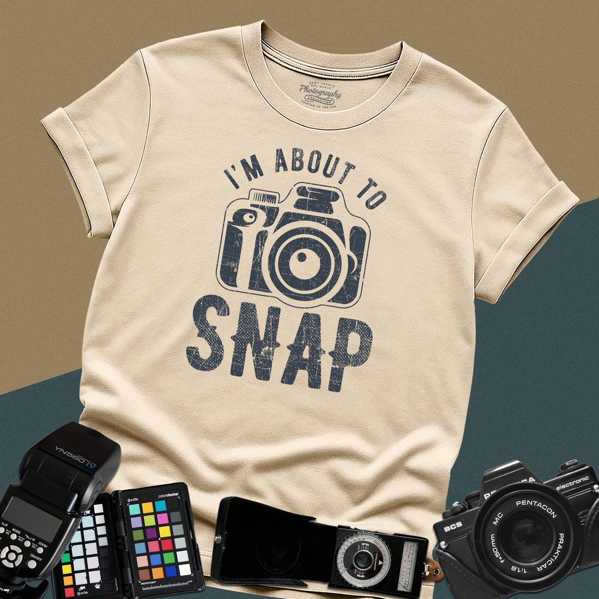 A sand t-shirt featuring a distressed modern camera graphic centered between the text "I'm about to" in a curved font above and "SNAP" written in large block letters below.