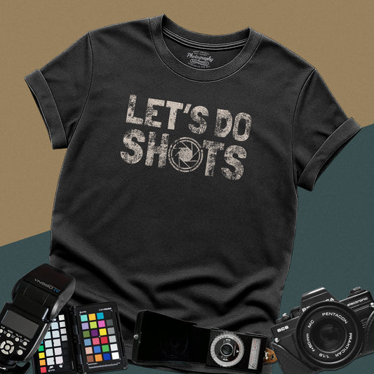 Heavy distressed black t-shirt featuring bold retro typography text "Let's Do Shots" with a camera aperture lens icon inside the letter O.