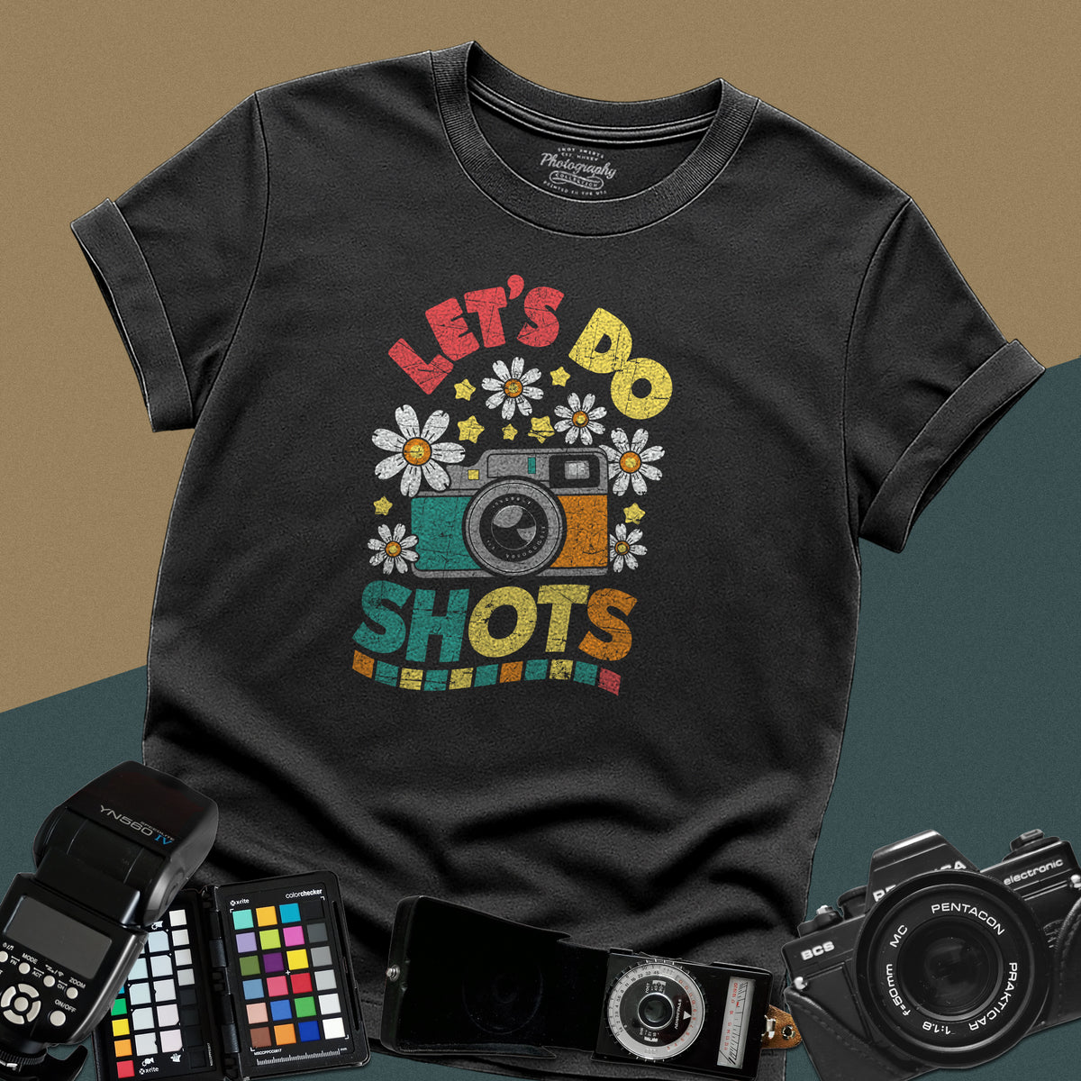 A black t-shirt featuring a colorful distressed retro graphic with a camera, daisies, and stars between the text "LET'S DO" and "SHOTS" in bold block letters.