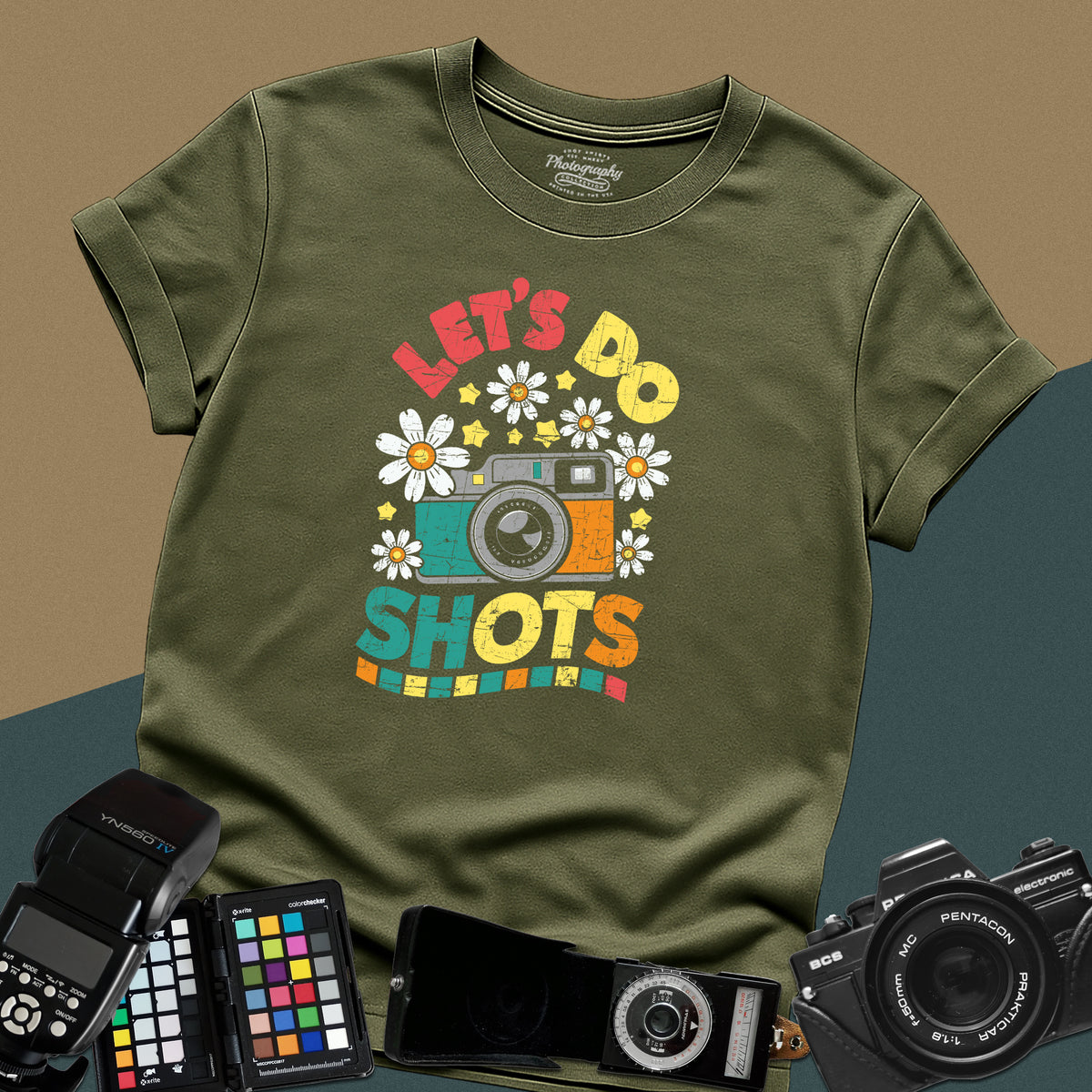 A military green t-shirt featuring a colorful distressed retro graphic with a camera, daisies, and stars between the text "LET'S DO" and "SHOTS" in bold block letters.