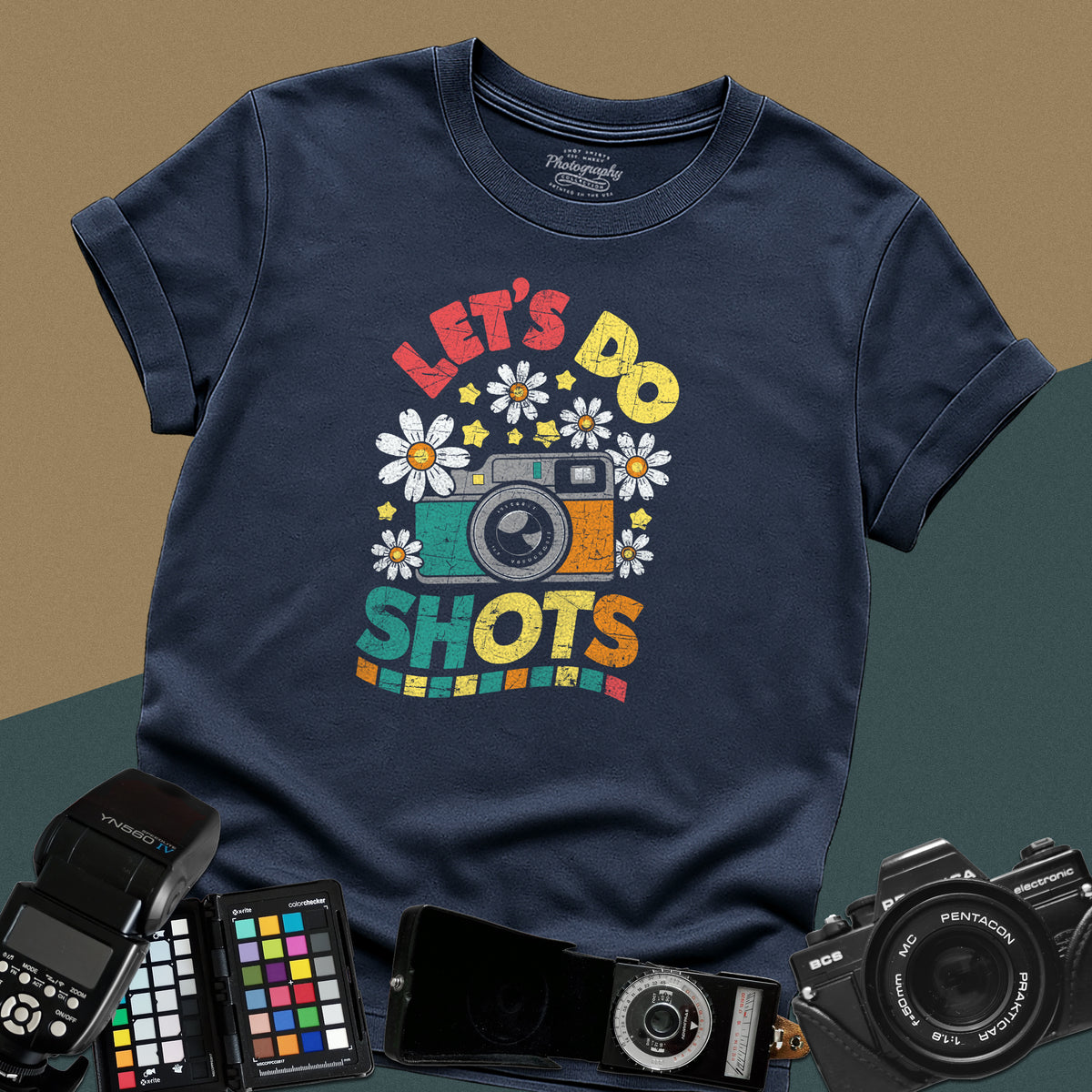 A navy blue t-shirt featuring a colorful distressed retro graphic with a camera, daisies, and stars between the text "LET'S DO" and "SHOTS" in bold block letters.