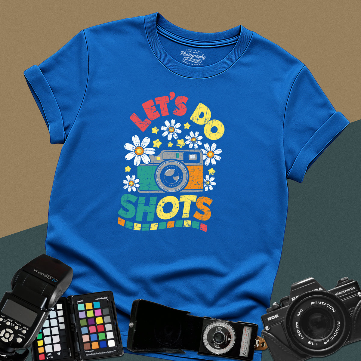 A royal blue t-shirt featuring a colorful distressed retro graphic with a camera, daisies, and stars between the text "LET'S DO" and "SHOTS" in bold block letters.