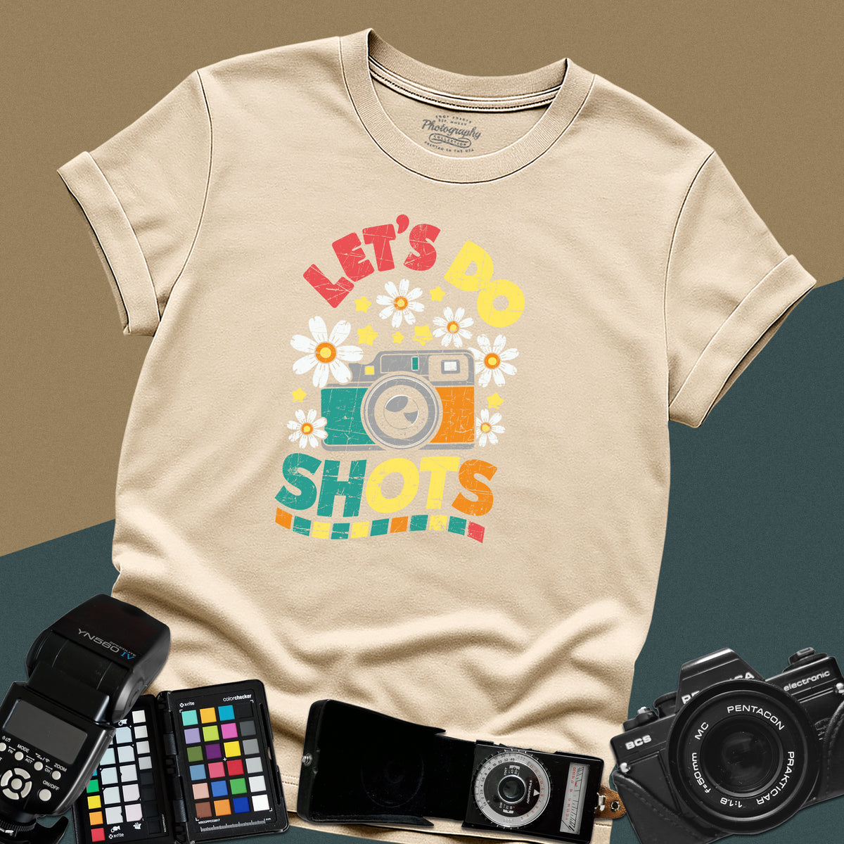 A sand t-shirt featuring a colorful distressed retro graphic with a camera, daisies, and stars between the text "LET'S DO" and "SHOTS" in bold block letters.