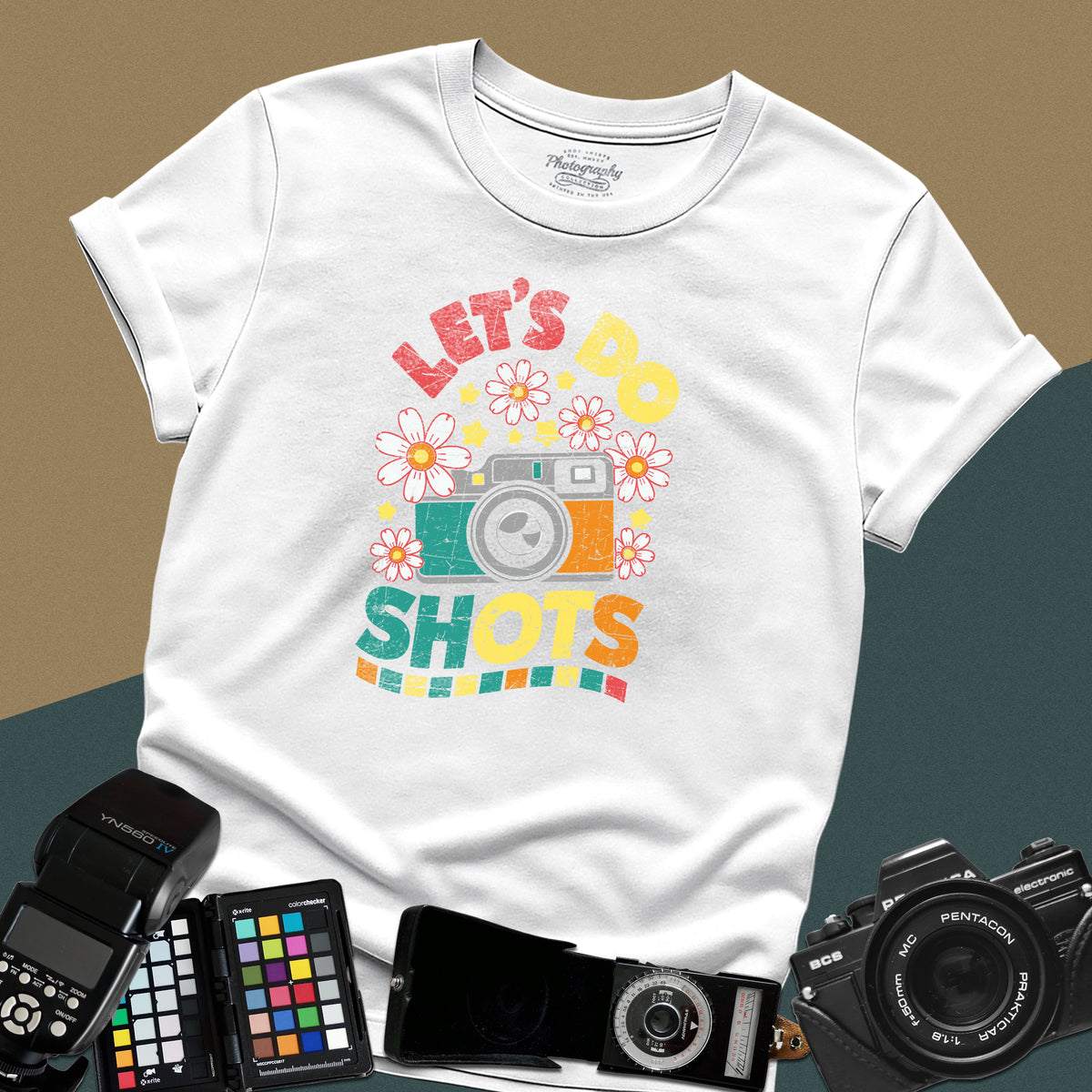 A white t-shirt featuring a colorful distressed retro graphic with a camera, daisies, and stars between the text "LET'S DO" and "SHOTS" in bold block letters.