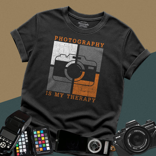 A casual black t-shirt featuring a unique graphic of a camera split into four textured squares with orange retro accents and the phrase "Photography Is My Therapy" written in a vintage distressed font.